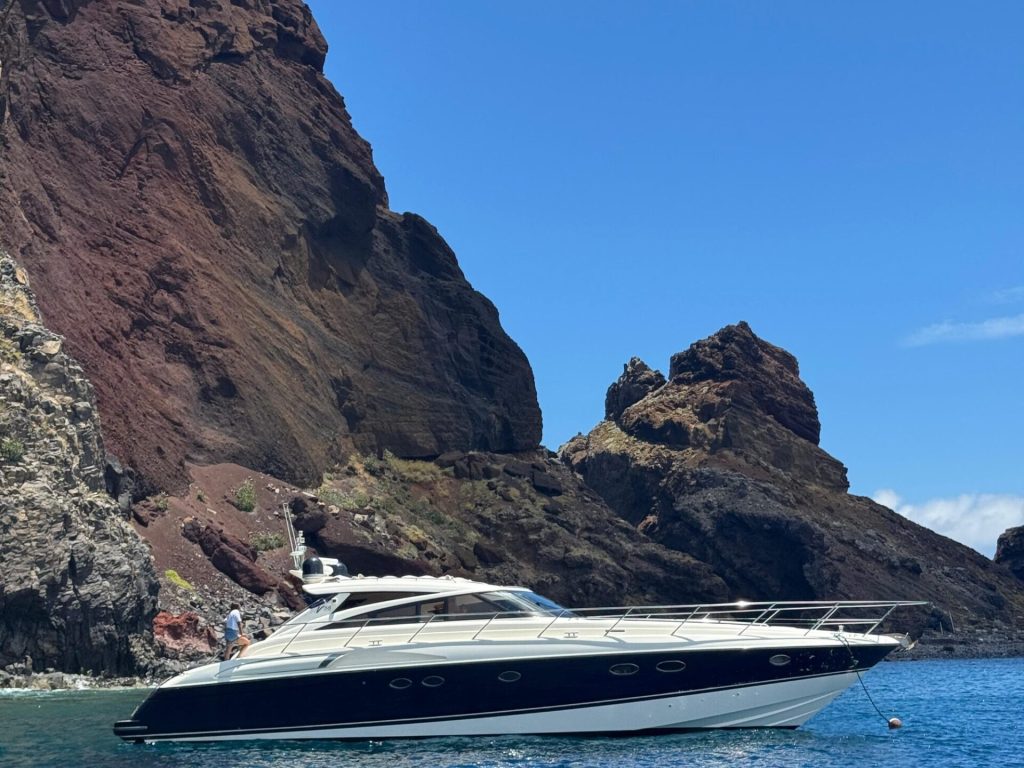 A sleek white and navy yacht floats on clear blue water at the base of towering, rugged volcanic cliffs under a bright, cloudless sky, an experience in a Luxury Yacht Charter near Desertas Island