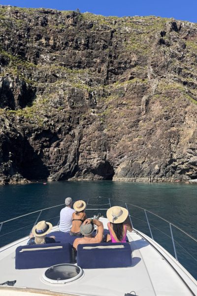 A group of friends lounges on the bow of a yacht, gazing up at towering volcanic cliffs rising straight from the calm, clear Atlantic water, an experience in a Luxury Yacht Charter near Desertas Island