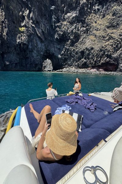 A group of friends relaxes on the cushioned bow of a boat, sunbathing and taking photos in calm turquoise water beneath towering volcanic cliffs and a small sea cave near Ponta de São Lourenço, Madeira,An experience in a Luxury Yacht Charter experience.