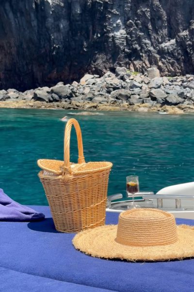 A straw hat, wicker basket, and drink rest on deep blue cushions at the bow of a boat, with crystal-clear turquoise water and rugged volcanic rocks rising steeply in the background. an experience by Luxury Charter Madeira.