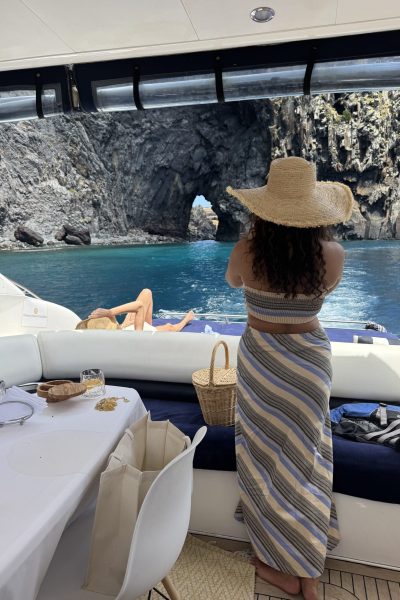 A woman in a striped dress and wide straw hat stands on the deck of a yacht, gazing at a dramatic rock arch rising from the turquoise sea near Ponta de São Lourenço in Madeira. an experience by Luxury Charter Madeira.