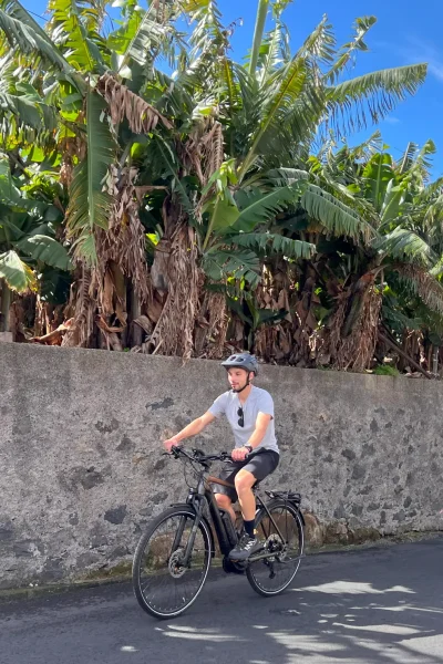 Two people riding e-bikes on a sunny road beside tall green banana trees, enjoying an E-Bike Road Tour, an experience by MADcycle