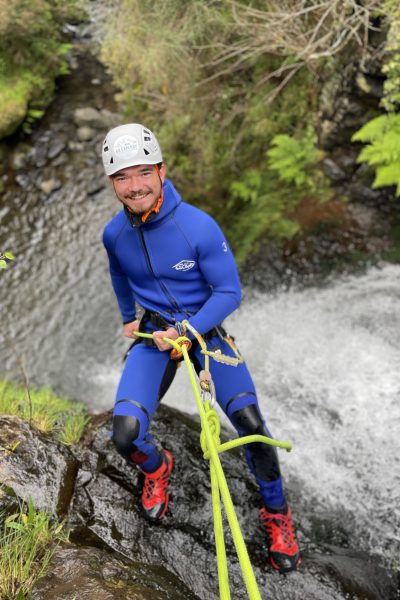 A smiling male canyoneer stands poised on a ledge, ready to descend a fern-surrounded stream drop in Porto Novo, Camacha, during a Beginner Level 1 tour with Go Explore Madeira