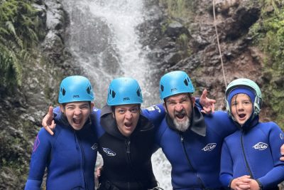 A joyful family of four canyoneers poses arm-in-arm in stream waters with an instructor rappelling a waterfall above in Porto Novo, Camacha, during a Beginner Level 1 tour with Go Explore Madeira.