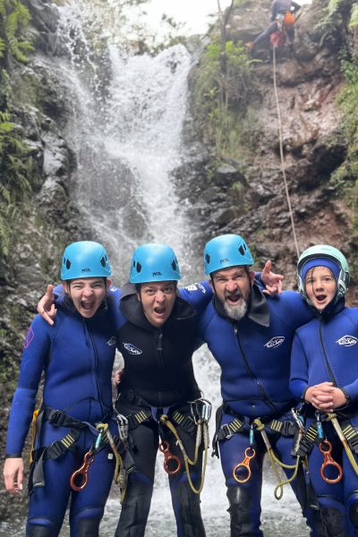 A joyful family of four canyoneers poses arm-in-arm in stream waters with an instructor rappelling a waterfall above in Porto Novo, Camacha, during a Beginner Level 1 tour with Go Explore Madeira.