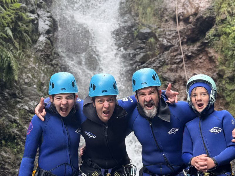 A joyful family of four canyoneers poses arm-in-arm in stream waters with an instructor rappelling a waterfall above in Porto Novo, Camacha, during a Beginner Level 1 tour with Go Explore Madeira.