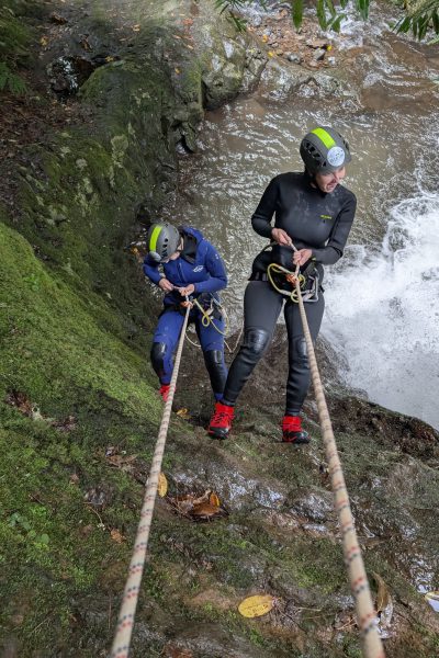 Two female canyoneers rappelling down a mossy rock face beside a stream in Porto Novo, Camacha, during a Beginner Level 1 canyoning tour with Go Explore Madeira.