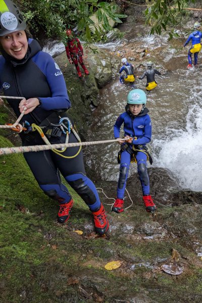 A female canyoneer helps a young child rappeller while the group watches in Porto Novo, Camacha, during a Beginner Level 1 canyoning tour with Go Explore Madeira.