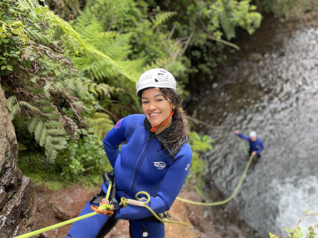 A smiling female canyoneer stands poised on a ledge, ready to descend a fern-surrounded stream drop in Porto Novo, Camacha, during a Beginner Level 1 tour with Go Explore Madeira.