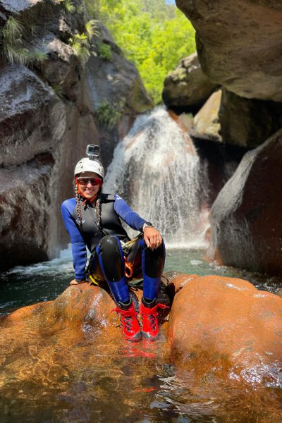 a female canyoneer relaxes on a rock beside a small waterfall in Ribeira do Cidrão, Nuns Valley, during an Intermediate Level 2 canyoning session.