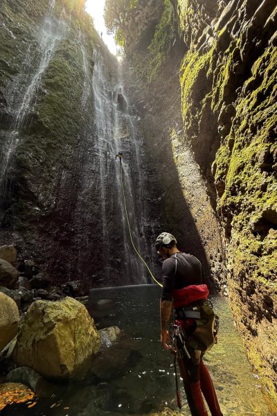 A guide watches as a canyoneer abseils a towering sunlit waterfall in Ribeira Funda Inferior during an Advanced Level 3 canyoning experience with Go Explore Madeira.