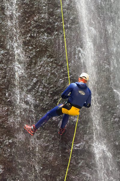 A canyoning guide in a blue and yellow wetsuit and helmet rappels down the vertical rock wall of Ribeira Funda Inferior’s main waterfall on a bright yellow rope during an Advanced Level 3 canyoning experience with Go Explore Madeira.