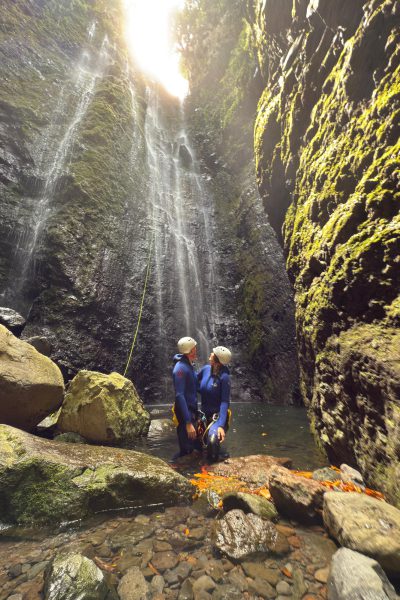 Two canyoneers in blue wetsuits and helmets stand hand-in-hand at the base of dual cascading waterfalls in a narrow moss-covered gorge, illuminated by dramatic sunlight rays piercing the canopy above a rocky pool with scattered boulders in Madeira's laurel forest during an Advanced Level 3 canyoning experience with Go Explore Madeira.