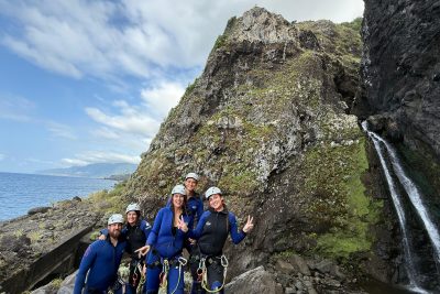 A group of five canyoneers in blue wetsuits and helmets pose triumphantly with peace signs and thumbs up beside a thin waterfall cascading from mossy volcanic cliffs into the Atlantic Ocean, with distant Desertas Islands and clouds on the horizon in Madeira during an Advanced Level 3 canyoning experience with Go Explore Madeira.