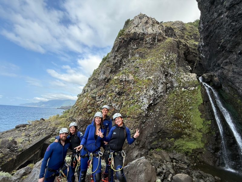 A group of five canyoneers in blue wetsuits and helmets pose triumphantly with peace signs and thumbs up beside a thin waterfall cascading from mossy volcanic cliffs into the Atlantic Ocean, with distant Desertas Islands and clouds on the horizon in Madeira during an Advanced Level 3 canyoning experience with Go Explore Madeira.