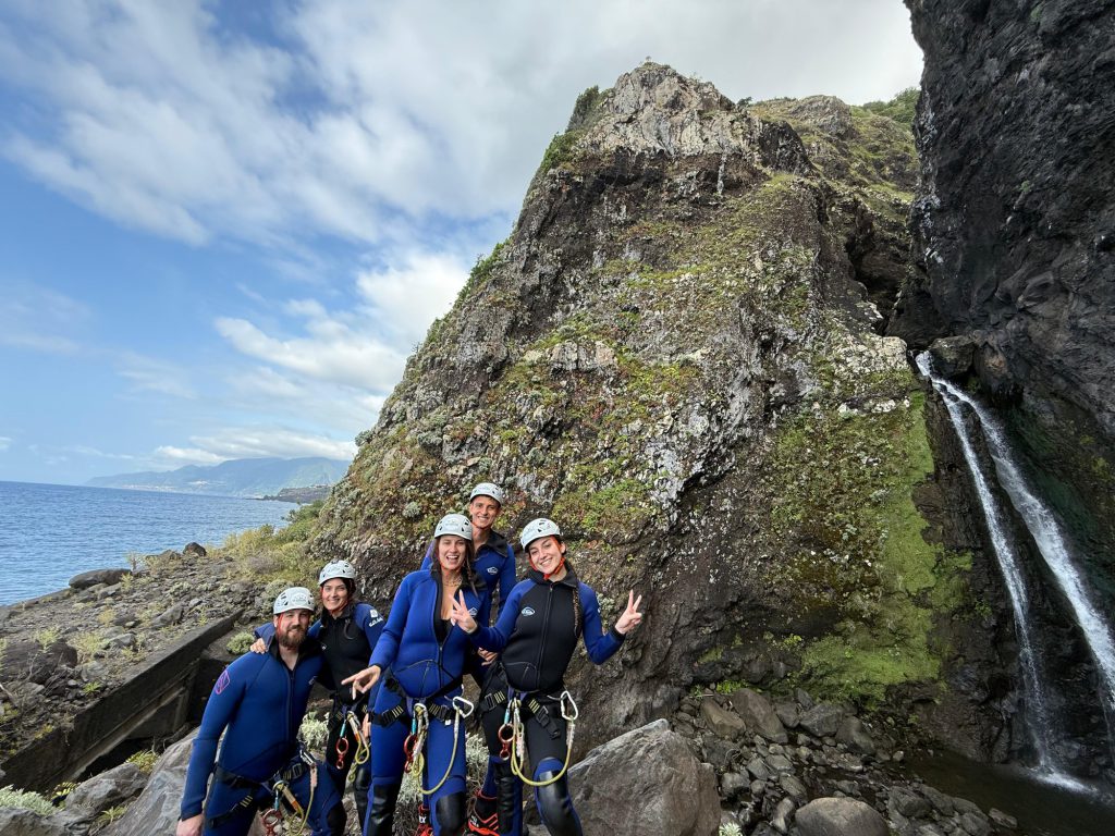 A group of five canyoneers in blue wetsuits and helmets pose triumphantly with peace signs and thumbs up beside a thin waterfall cascading from mossy volcanic cliffs into the Atlantic Ocean, with distant Desertas Islands and clouds on the horizon in Madeira during an Advanced Level 3 canyoning experience with Go Explore Madeira.