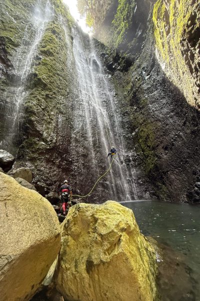 Two canyoneers in red gear rappel down a mossy rock face beside a tall cascading waterfall in the dramatic Ribeira Funda canyon on Madeira's north coast, surrounded by sheer green volcanic walls, large yellow boulders, and a clear pool below under beams of sunlight during an Advanced Level 3 canyoning experience with Go Explore Madeira.