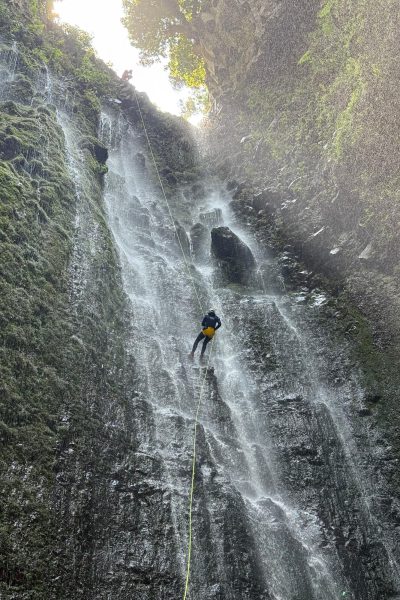 A canyoneer abseils down a towering sunlit waterfall in Ribeira Funda Inferior Inferior during an Advanced Level 3 canyoning experience with Go Explore Madeira.