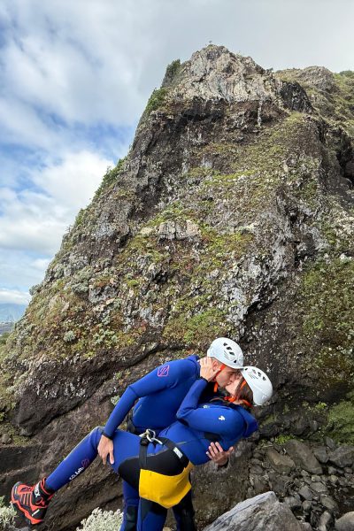 A couple kissing in blue wetsuits and helmets beside a thin waterfall cascading from mossy volcanic cliffs into the Atlantic Ocean, with distant Desertas Islands and clouds on the horizon in Madeira during an Advanced Level 3 canyoning experience with Go Explore Madeira.