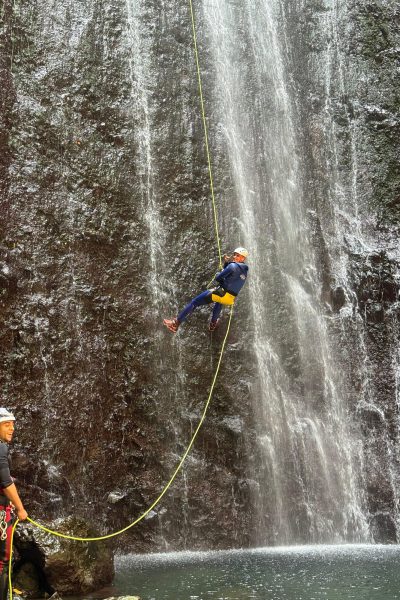 A guide watches as a canyoneer abseils a towering sunlit waterfall in Ribeira Funda Inferior during an Advanced Level 3 canyoning experience with Go Explore Madeira.
