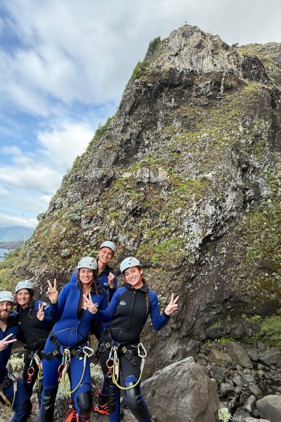 A group of five canyoneers in blue wetsuits and helmets pose triumphantly with peace signs and thumbs up beside a thin waterfall cascading from mossy volcanic cliffs into the Atlantic Ocean, with distant Desertas Islands and clouds on the horizon in Madeira during an Advanced Level 3 canyoning experience with Go Explore Madeira.