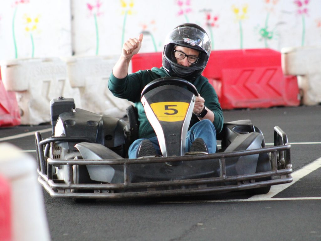 Indoor go‑kart driver in kart number 5 giving a thumbs-up while racing on a track located under the Madeira Airport with safety barriers and colorful wall decorations in the background.An experience by Go Kart Madeira