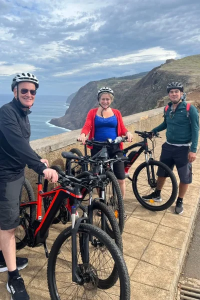 Three riders posing with their e-bikes at a dramatic cliffside viewpoint above the ocean on the Bike tour on the west coast of Madeira, an experience by Madeira Island Route.
