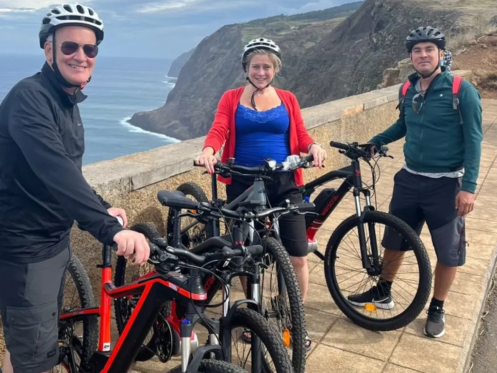 Three riders posing with their e-bikes at a dramatic cliffside viewpoint above the ocean on the Bike tour on the west coast of Madeira, an experience by Madeira Island Route.