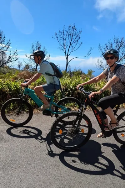 Two riders pedaling e-bikes along a sunny coastal road lined with greenery and ocean views on the Bike tour on the west coast of Madeira, an experience by Madeira Island Route.