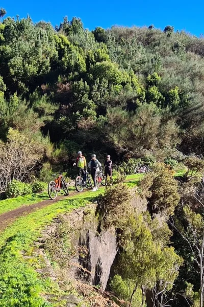 Small group of riders pausing with their e-bikes on a narrow levada trail above a lush green valley on the Levada da Camacha Trail by E-Bike, an experience by Madeira Island Route.