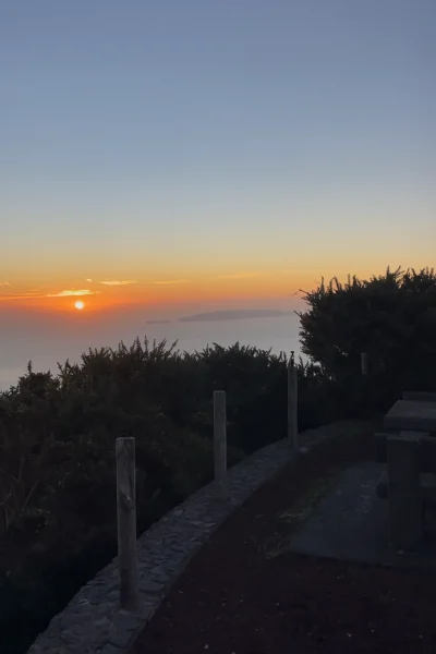 Sunset view from a clifftop picnic area with stone tables, low bushes and wooden posts along a stone path, overlooking the ocean and a distant island under a clear sky. A Jeep tour experience by Sea La Vie Mountain Expedition.