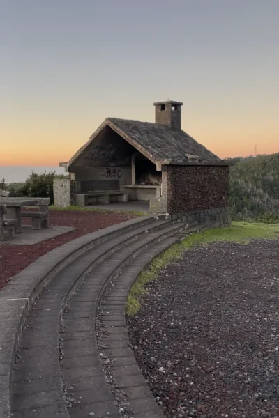 Stone picnic shelter with chimney and benches at a scenic hillside viewpoint, curved paved steps and gravel foreground, under a clear pastel sunset sky. An experience by Sea La Vie Mountain Expedition.