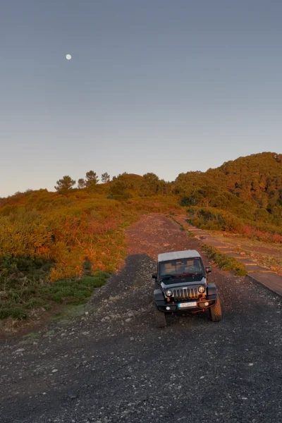 Sunset off‑road scene with a black Jeep Wrangler parked on a rocky gravel track, surrounded by green and orange-lit hillsides, under a clear sky with the moon visible above.An experience by Sea La Vie Mountain Expedition.