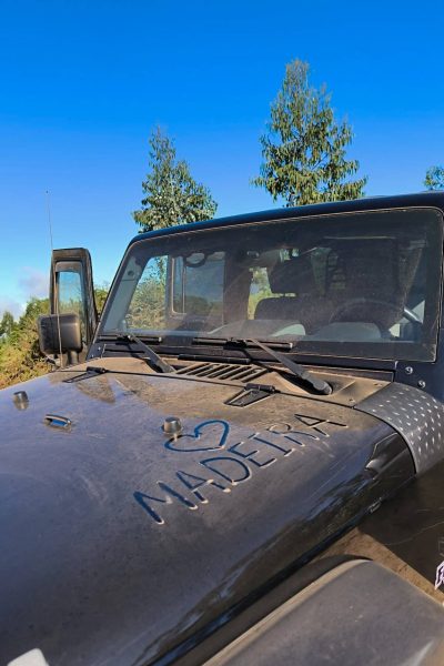 Close-up of a dusty black Jeep hood with a "Heart Madeira” drawn in the dirt, set against a clear blue sky and tall green trees in the background, during an offroad tour by Sea La Vie Mountain Expedition.