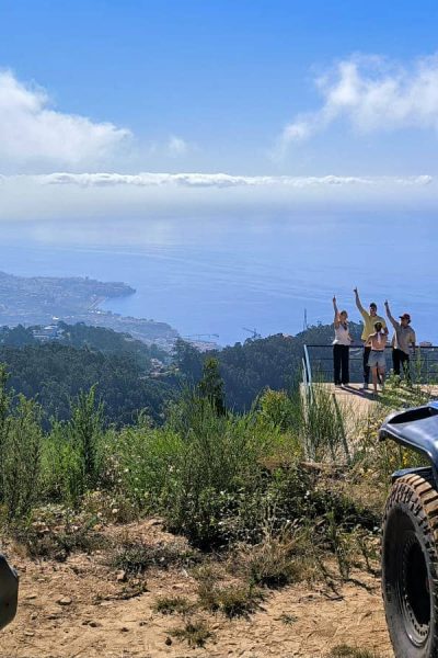 Panoramic hilltop viewpoint in Madeira with two Jeeps parked in the foreground, a woman filming on the left, and a group raising their arms on a distant platform overlooking the coastline and Atlantic Ocean., during an offroad tour by Sea La Vie Mountain Expedition.