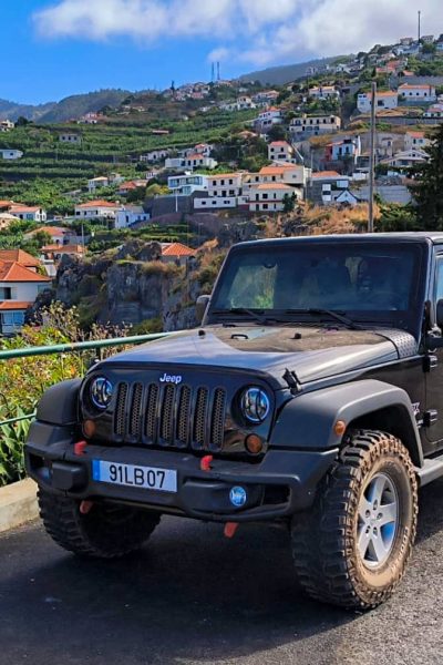 Black Jeep Wrangler parked at a roadside viewpoint in Madeira, with a person taking photos over a green railing and a hillside village of terraced houses and mountains in the background. A Jeep tour experience by Sea La Vie Mountain Expedition.