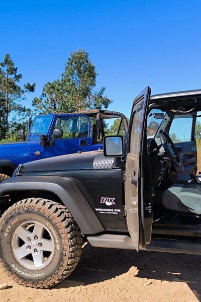Group of friends chatting beside two off-road Jeeps on a sunny dirt clearing in Madeira, with the doors of a black Wrangler open and a blue Jeep parked in front, surrounded by tall green trees. A Jeep tour experience by Sea La Vie Mountain Expedition.