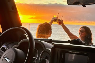 Interior view from a Jeep looking out toward the ocean at sunset, with two people outside clinking drinks against a vibrant orange and yellow sky over the water.A Jeep tour experience by Sea La Vie Mountain Expedition.