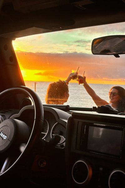 Interior view from a Jeep looking out toward the ocean at sunset, with two people outside clinking drinks against a vibrant orange and yellow sky over the water.A Jeep tour experience by Sea La Vie Mountain Expedition.