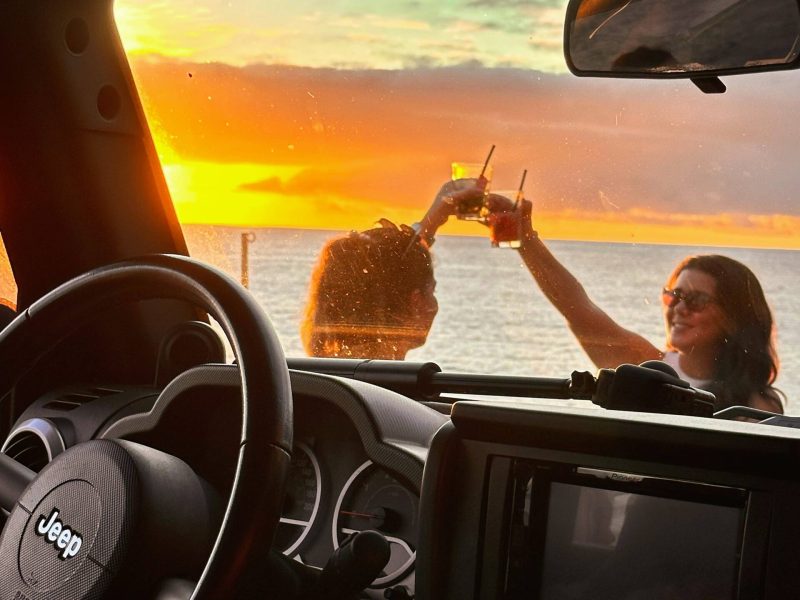 Interior view from a Jeep looking out toward the ocean at sunset, with two people outside clinking drinks against a vibrant orange and yellow sky over the water.A Jeep tour experience by Sea La Vie Mountain Expedition.