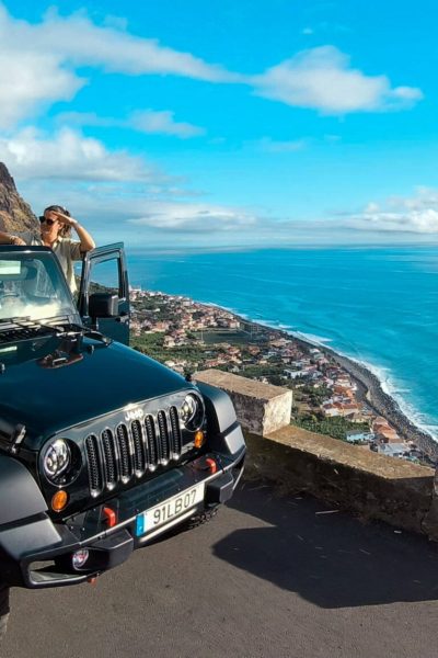 Family enjoying a scenic stop with a black Jeep Wrangler at a cliffside viewpoint in Madeira, doors open and arms raised, overlooking a coastal town and the bright blue Atlantic Ocean. A Jeep tour experience by Sea La Vie Mountain Expedition.