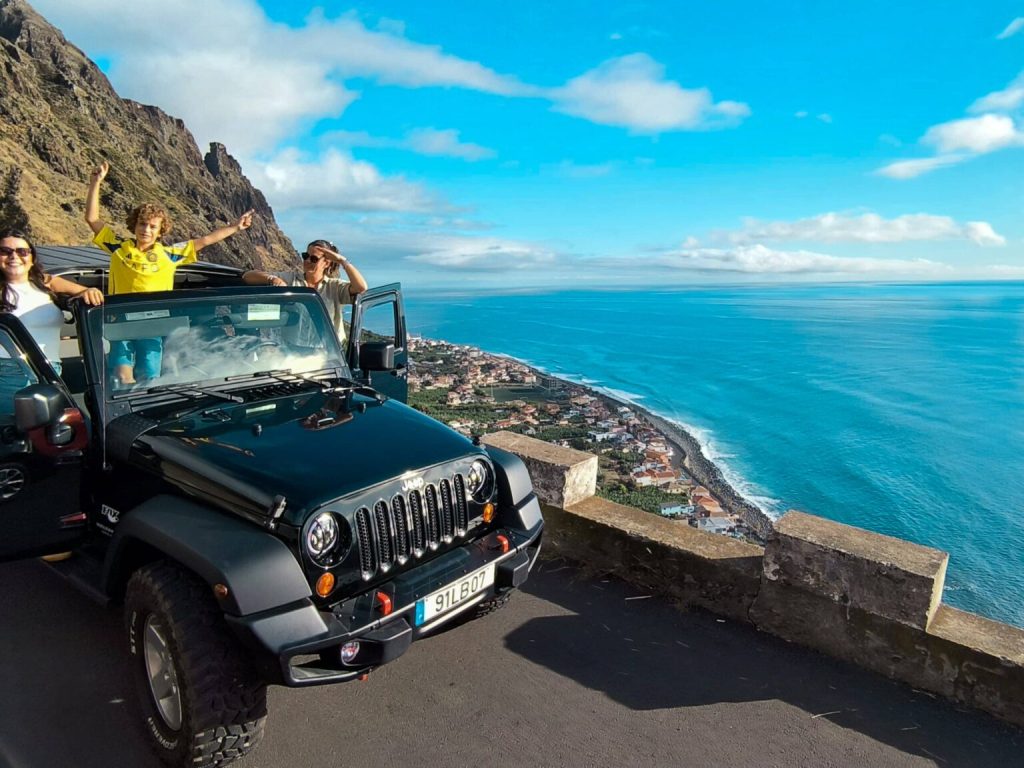 Family enjoying a scenic stop with a black Jeep Wrangler at a cliffside viewpoint in Madeira, doors open and arms raised, overlooking a coastal town and the bright blue Atlantic Ocean. A Jeep tour experience by Sea La Vie Mountain Expedition.