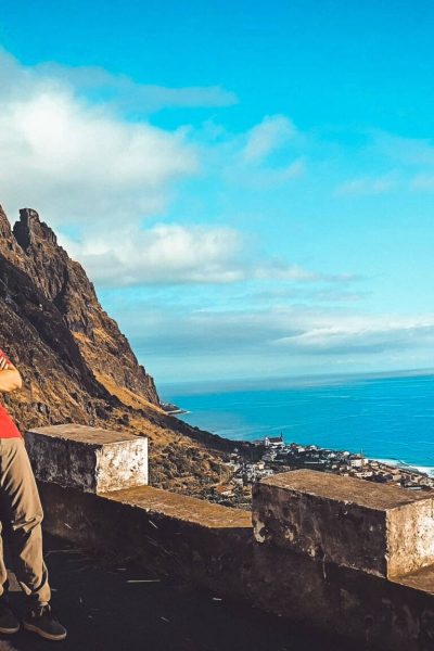 Couple standing by a black Jeep Wrangler at a scenic cliffside viewpoint in Madeira, overlooking a coastal town, steep rocky mountains and the turquoise Atlantic Ocean under a bright blue sky. Madeira North & West Mountain Jeep Expedition Tour Adventure by Sea La Vie Mountain Expedition.