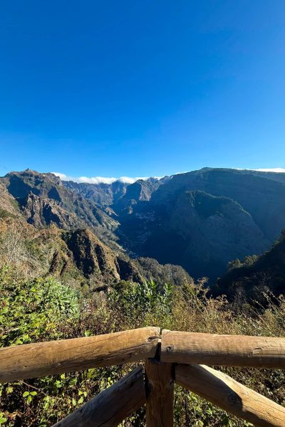 Mountain viewpoint in Madeira with a wooden railing in the foreground, overlooking a deep green valley surrounded by rugged peaks under a clear, bright blue sky. A Jeep tour experience by Sea La Vie Mountain Expedition.