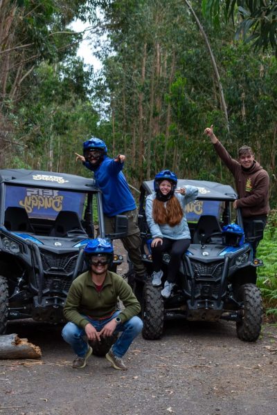 Seven thrilled adventurers in helmets strike celebratory poses beside two black Let’s Buggy Can-Am Mavericks parked on a forest trail deep within Madeira’s UNESCO-listed Laurissilva rainforest, during a Let's Buggy's Happy Hour Tour experience in Madeira.