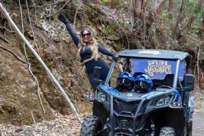 A Happy blonde woman in sunglasses and black athletic wear poses triumphantly beside a blue Let’s Buggy Can-Am Maverick buggy parked on a rocky trail in Madeira’s Laurissilva forest, with two helmets on the seats, during a Let's Buggy's Happy Hour Tour experience in Madeira.