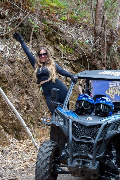 A Happy blonde woman in sunglasses and black athletic wear poses triumphantly beside a blue Let’s Buggy Can-Am Maverick buggy parked on a rocky trail in Madeira’s Laurissilva forest, with two helmets on the seats, during a Let's Buggy's Happy Hour Tour experience in Madeira.