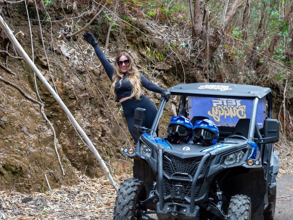 A Happy blonde woman in sunglasses and black athletic wear poses triumphantly beside a blue Let’s Buggy Can-Am Maverick buggy parked on a rocky trail in Madeira’s Laurissilva forest, with two helmets on the seats, during a Let's Buggy's Happy Hour Tour experience in Madeira.