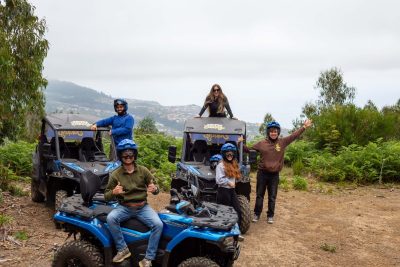 A group of six excited participants in helmets pose with thumbs up beside two blue Can-Am buggies from Let’s Buggy, overlooking misty hills and distant mountains in Madeira’s highlands, during a Let's Buggy's Laurissilva experience in Madeira.