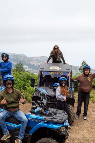 A group of six excited participants in helmets pose with thumbs up beside two blue Can-Am buggies from Let’s Buggy, overlooking misty hills and distant mountains in Madeira’s highlands, during a Let's Buggy's Laurissilva experience in Madeira.